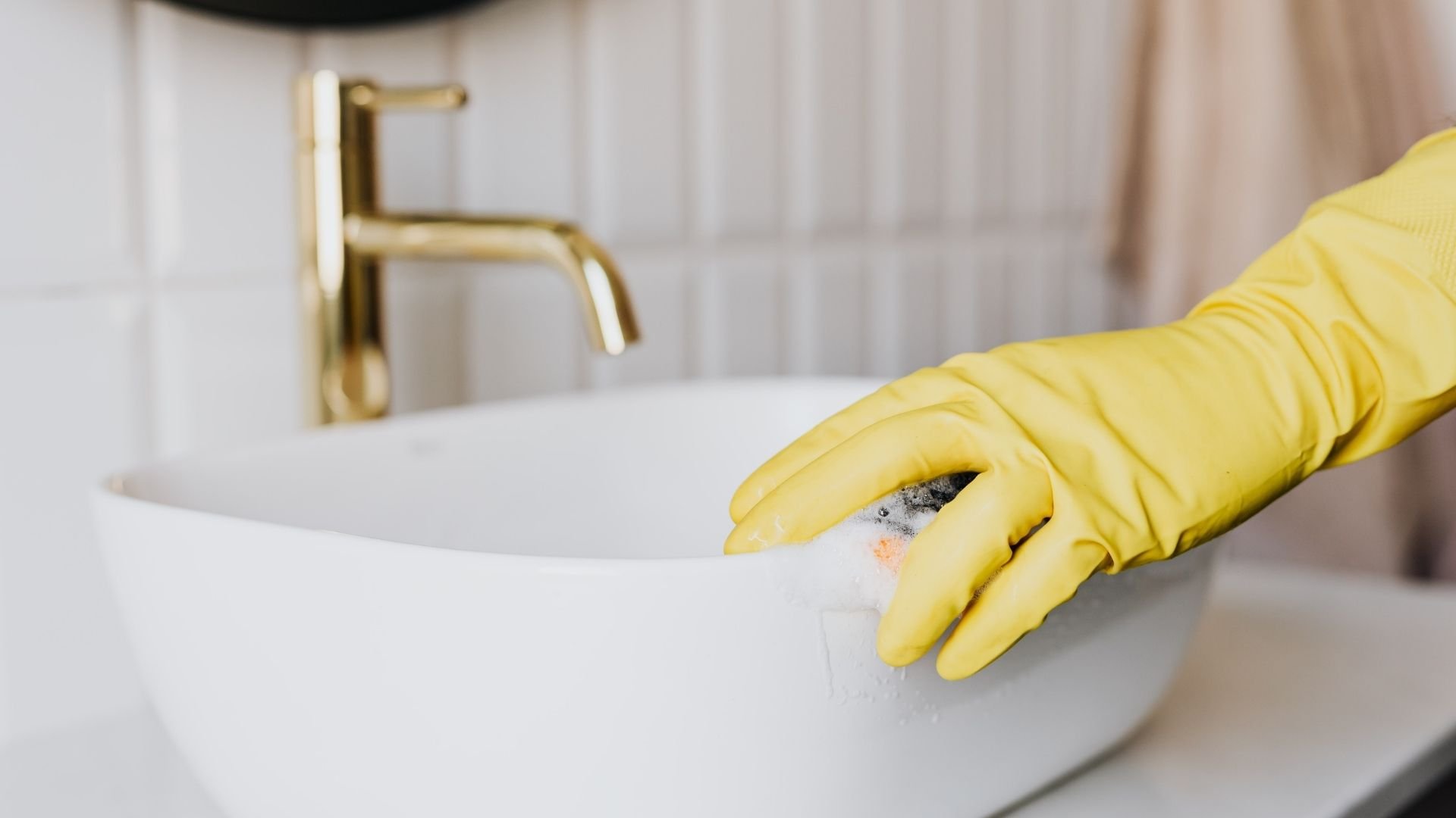 Yellow gloved hand cleaning white sink with gold faucet